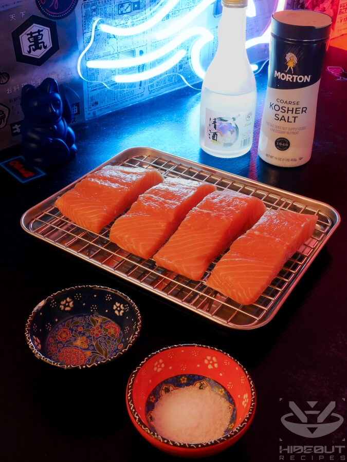 Four salmon fillets are laid out on a metal rack over a drip pan. Small bowls containing pre-portioned sake and kosher salt are in the foreground.