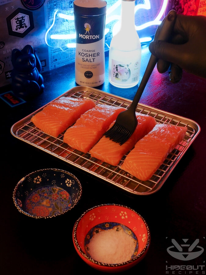 A gloved hand is using a silicone brush to apply sake to four salmon fillets on a metal rack over a drip pan.