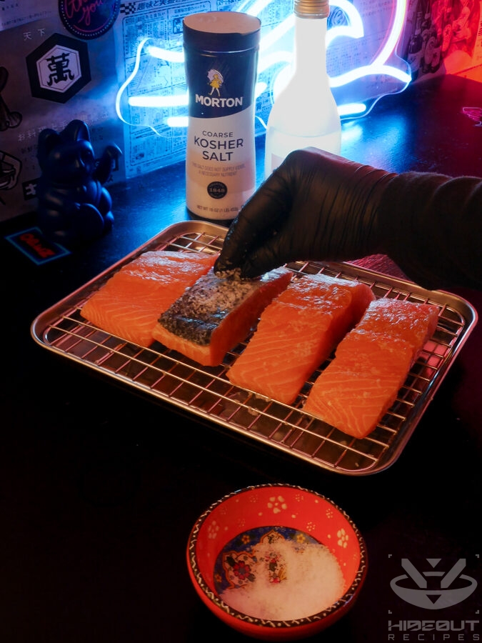 A gloved hand is rubbing coarse kosher salt on four salmon fillets that are placed on a metal rack over a drip pan.