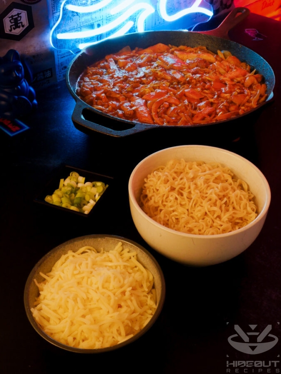 A large cast-iron pan of Korean Dakgalbi is surrounded by smaller bowls filled with scallions, grated cheese, and pre-cooked instant noodles.