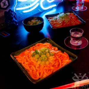 A futuristic Cyberpunk-style food photo showing a plate of Gochujang Udon. Glowing chopsticks, a bowl of scallions, and a glass of soju surround it.