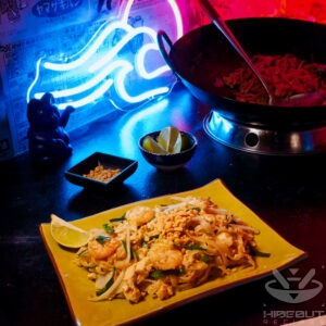 A futuristic Cyberpunk-style food photo showing a plate of Pad Thai in the foreground. Small bowls with toppings, glowing chopsticks, and a large wok surround it.