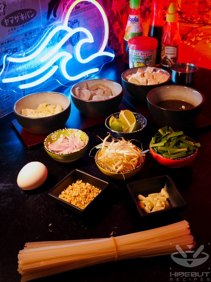 Pre-measured and chopped Pad Thai ingredients and toppings are distributed to small bowls on a table, plus some uncooked noodles and condiments.