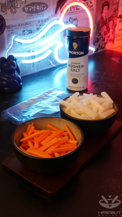 Julienned carrots and radish are placed in two small bowls, with coarse kosher salt and an empty freezer bag behind them.