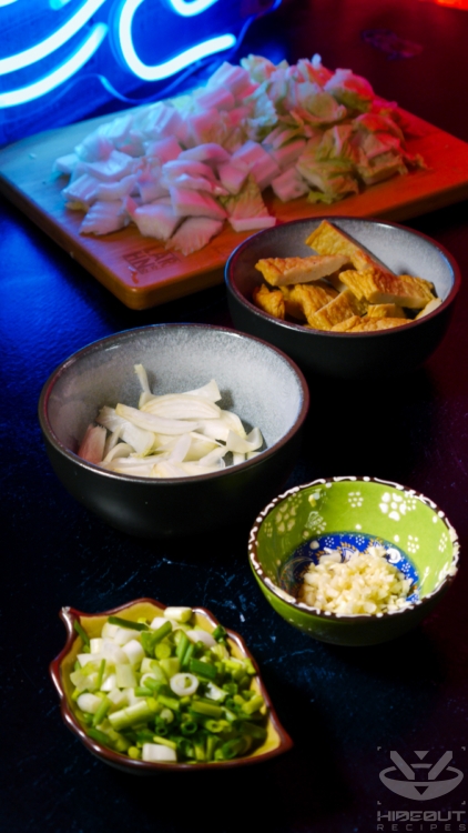 Raw, chopped up ingredients for Rabokki are distributed to small bowls: scallions, garlic, shallot, Korean rice cakes, and napa cabbage.