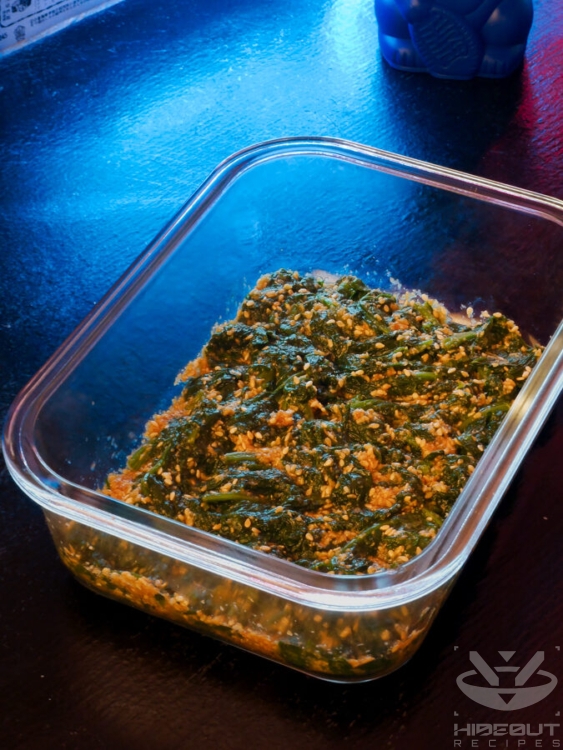 Cooked spinach is mixed with sesame seasoning inside of a glass Tupperware container for Japanese Sesame Spinach.