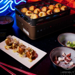Some Takoyaki on a plate with toppings. More Takoyaki can be seen being made on a cassette in the background.
