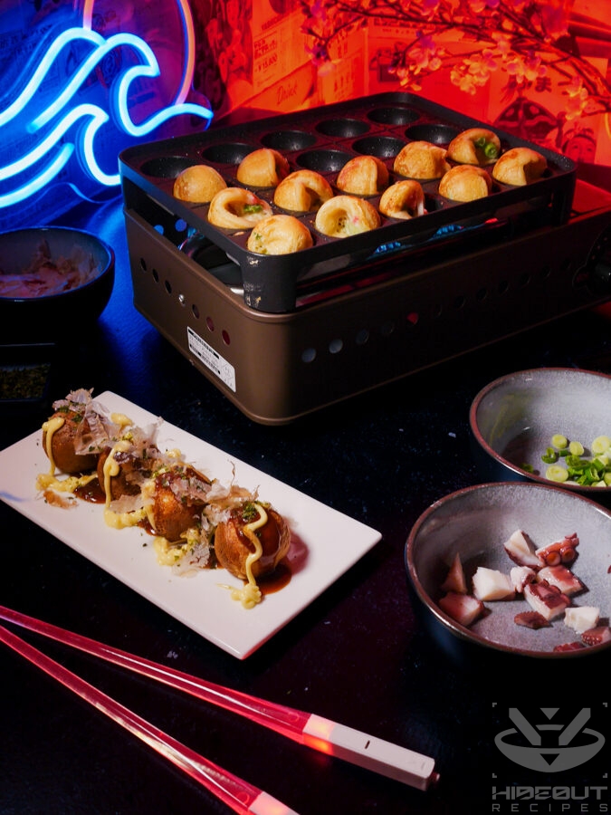 Some Takoyaki on a plate with toppings. More Takoyaki can be seen being made on a small grill in the background.