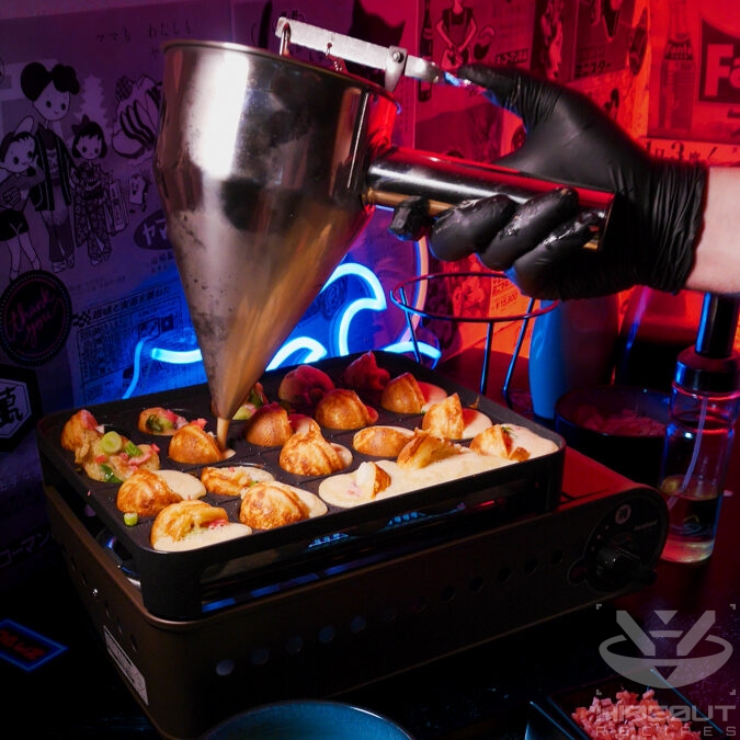 A Takoyaki grill whose holes are filled with batter and fillings. A gloved hand is adding more dough using a dough dispenser.