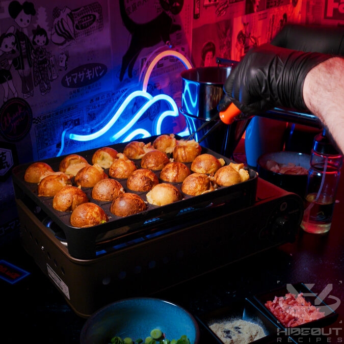 A specialized grill filled with Takoyaki balls being prepared. Gloved hands are using plastic picks to flip them around.