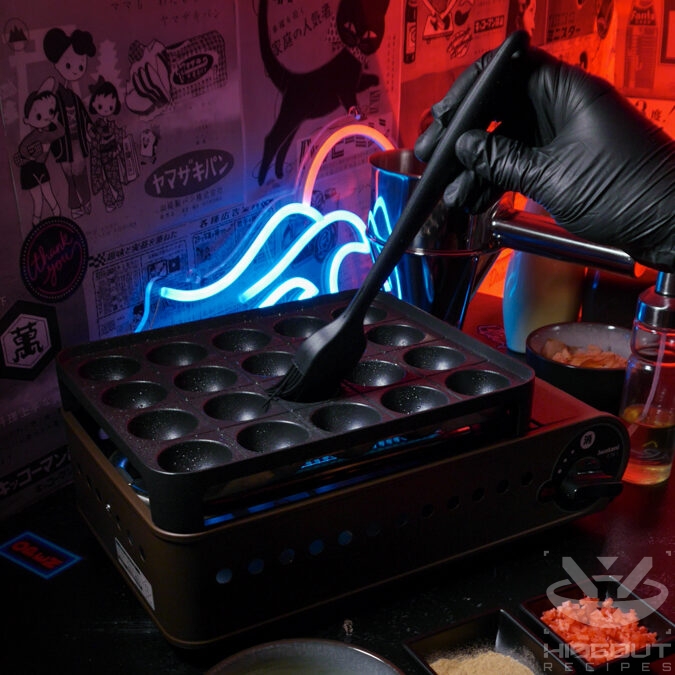 A gloved hand is brushing oil on a Takoyaki grill, prepared ingredients surrounding it.
