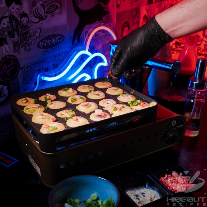 A Takoyaki grill whose holes are filled with batter. A gloved hand is sprinkling fillings into the dough.