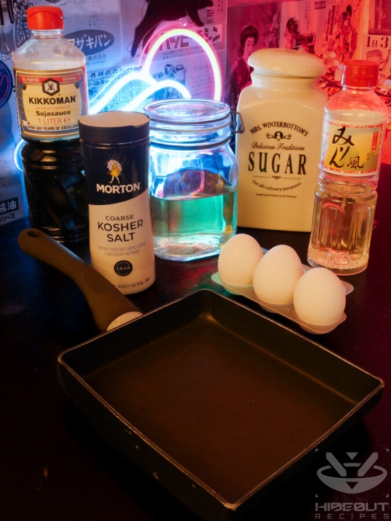 Ingredients for Tamagoyaki are gathered on the table in their packaging. Three eggs and a specialized rectangular Tamagoyaki pan are in the foreground.