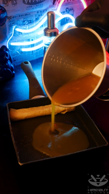 A gloved hand is pouring some pre-mixed Tamagoyaki from a measuring cup into an oiled rectangular pan. Some fried Tamagoyaki is already folded on one end of the pan.