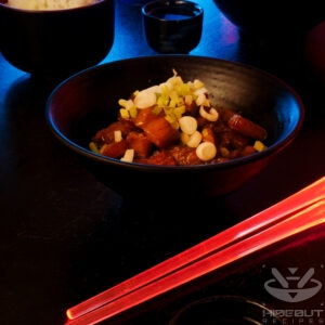 A futuristic Cyberpunk food photo. A bowl of oven-braised Kakuni (Japanese pork belly) is at the center, with a bowl of rice, chopsticks, and a cup of Sake around it.