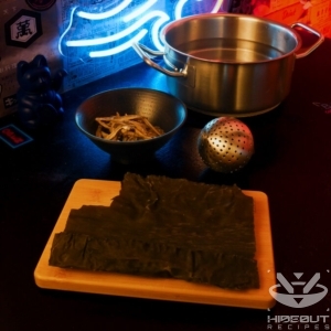 Ingredients for Korean soup stock are distributed on a table: kombu seaweed, anchovies, and a pot of water. A spice ball is lying nearby.