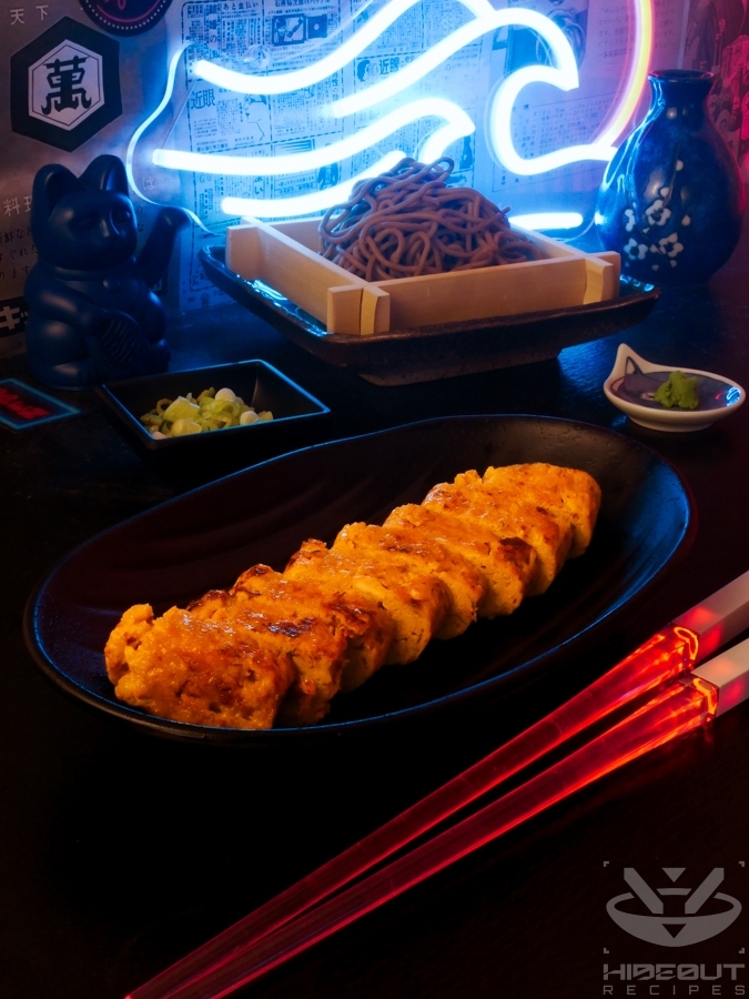 A futuristic Cyberpunk-style food photo of Tamagoyaki sliced into bite-sized pieces. Glowing chopsticks lie next to it, and a plate of cold soba can be seen in the background.