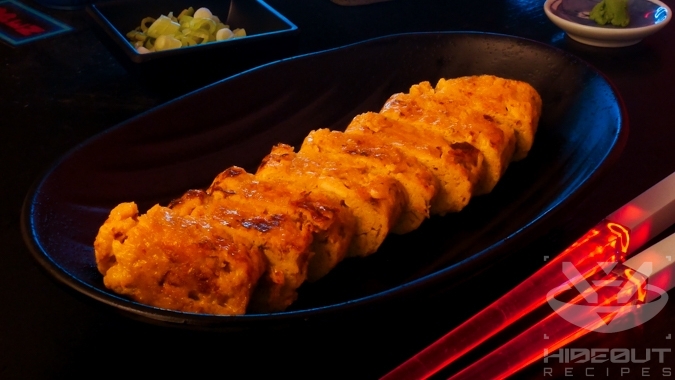 A futuristic Cyberpunk-style food photo showing a closeup of Tamagoyaki sliced into bite-sized pieces. Glowing chopsticks lie next to it.