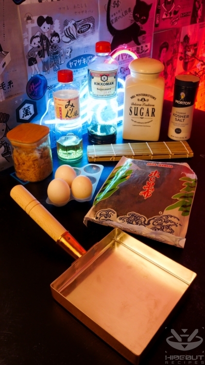 Ingredients for Tamagoyaki are gathered on the table in their packaging. Three eggs and a specialized rectangular Tamagoyaki pan are in the foreground.