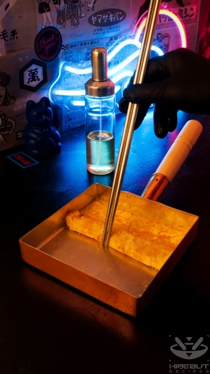 The fried layer of Tamagoyaki is being folded towards one side of the rectangular pan using long cooking chopsticks.