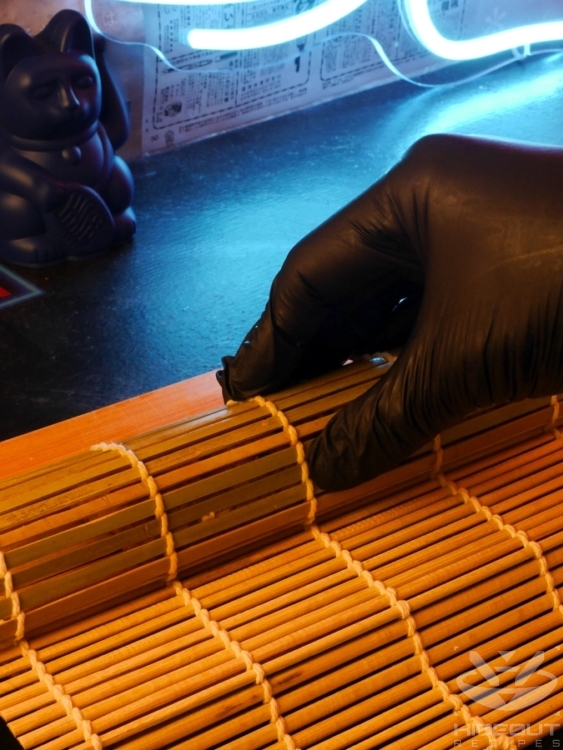 A gloved hand is using a small bamboo mat to press the Tamagoyaki into shape.