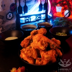 A futuristic Cyberpunk-style food photo. A pile of deep-fried Korean Fried Chicken is surrounded by Banchan and toppings. A rack holding silicone brushes with different sauces underneath can be seen in the background.
