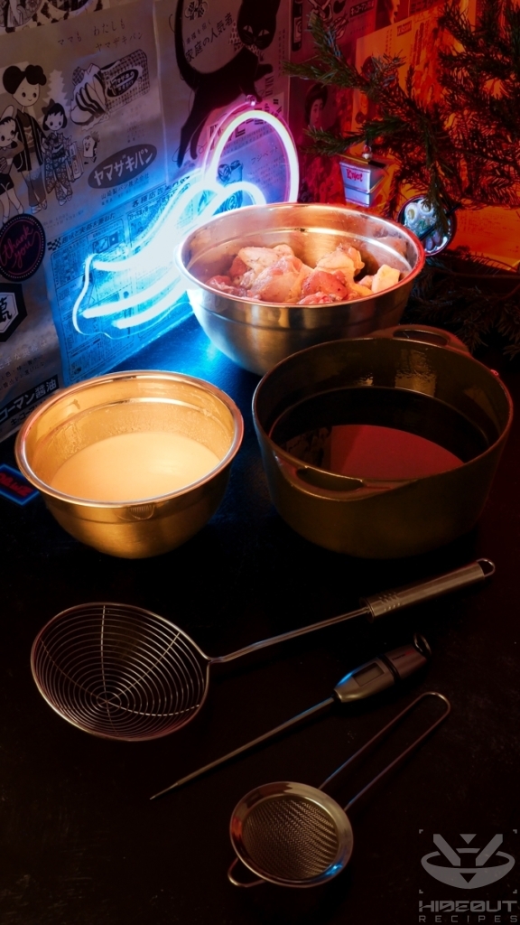 A bowl of dry-brined chicken, a bowl of mixed batter, and a cast iron pot of hot vegetable oil stand on a table. A spider skimmer, fine mesh skimmer, and thermometer lie in the foreground.