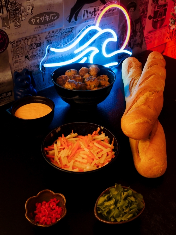 All prepared ingredients for bánh mì are laid out on a table, ready for final assembly: meatballs, Sriracha mayo, pickled radish and carrots, minced chili, chopped cilantro, and two large baguettes.