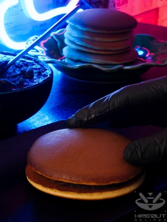 A hand closes a finished Dorayaki, consisting of two pancakes and red bean paste filling.